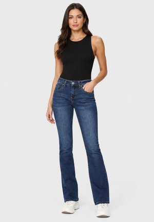 Frau steht in einem eng anliegenden schwarzen ärmellosen Top, blauen Bootcut-Jeans und weißen Sneakern vor einem schlichten hellen Hintergrund.