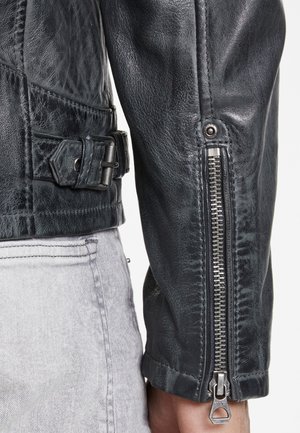 Schwarze Lederjacke mit strukturiertem Finish, Taillengurt und Reißverschlüssen an den Ärmeln. Detail des silbernen Reißverschlusses und der Nähte sichtbar.