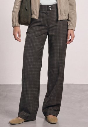 Femme portant un pantalon large à carreaux taille haute, une veste beige et des ballerines beiges, debout sur un sol gris lisse avec un fond clair.
