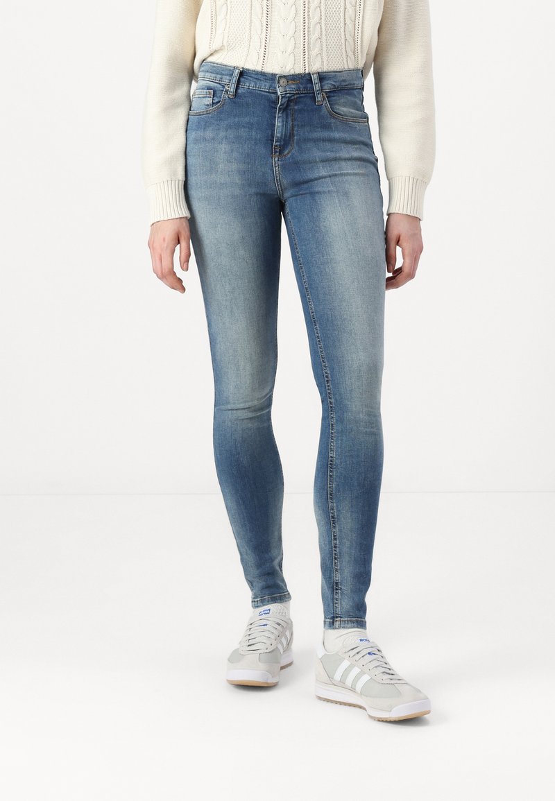 Fakó kék skinny farmernadrág sima textúrával, öt zsebes dizájnnal és övhurkokkal, krémszínű pulóverrel és fehér sportcipővel párosítva.