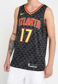 Man in een zwart Atlanta basketbalshirt met nummer 17, voorzien van rode en gele accenten, gecombineerd met witte shorts en zichtbare tatoeages op de linkerarm.