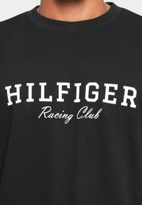 Čierna, textúrovaná mikina s výrazným bielym textom "HILFIGER" a kurzívou "Racing Club" nižšie. Okrúhly výstrih, neformálny dizajn.