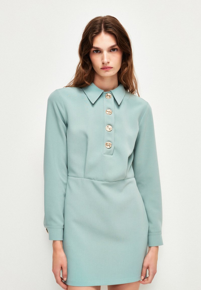 Vestito azzurro chiaro con maniche lunghe e colletto, dotato di cinque bottoni dorati sul davanti. Tessuto morbido con una silhouette aderente.