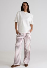 White cotton t-shirt with short sleeves and small logo, paired with wide striped pants in red and white, and brown footwear.