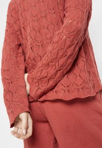 Jersey de punto en rojo apagado con un patrón de encaje abierto y texturizado, que presenta mangas largas y un dobladillo con forma de pico. Acompañado de pantalones a juego.