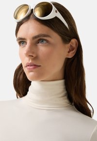 Hvid top med høj krave og en glat tekstur samt embossed logo "BOGNER", parret med runde guldsolbriller med hvide stel.