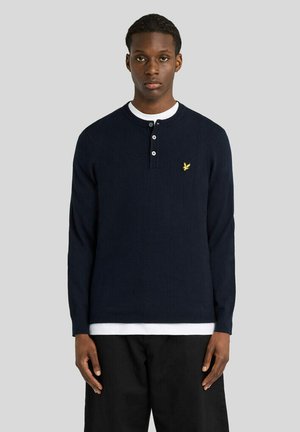 Jeune homme portant un t-shirt henley manches longues bleu marine avec des boutons blancs et un logo oiseau jaune sur la poitrine, debout devant un fond gris.