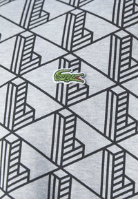 Gray fabric features a geometric black pattern with cubes and lines. A green embroidered crocodile logo is located at the bottom right.