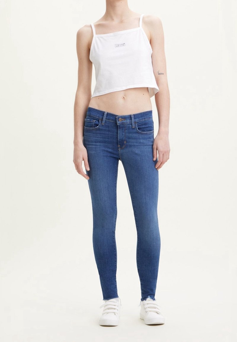 Top corto blanco sin mangas con tirantes finos y jeans ajustados azules. La modelo lleva zapatillas blancas. Denim suave y ajustado con un sencillo detalle de logo en la parte superior.