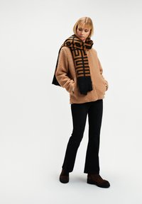 Kobieta w beżowym futrze teddy, czarnej wzorzystej szalu, czarnych spodniach i brązowych botkach, stojąca na tle jednolitego białego tła.