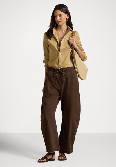 Žena oblečená v horčicovom tričku, voľných hnedých nohaviciach, v sandáloch a so svetlohnedou kabelkou cez rameno, stojaca pred jednofarebným pozadím.