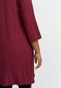 Burgundy tunika med vertikala texturerade ränder, trekvartsärmar och rundad nederkant. Lätt tyg med mjuk fall och subtil lyster.