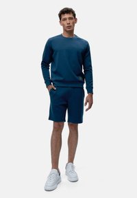 Temno modra pulover in kratke hlače, narejene iz gladkega materiala. Pulover ima krojen ovratnik; kratke hlače imajo žep in prostoren kroj. Bele teniske.