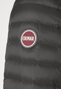 Näruppfattning av svart vadderat tyg med en cirkulär emblem-logotyp som har "COLMAR" i vitt mot en vinröd bakgrund.