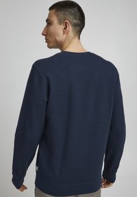 Tumši zila džemperis, teksturēta auduma, ar apaļu kakla izgriezumu, garām piedurknēm un ribotām malām. Minimāls zīmols sānos netālu no apakšmalas.