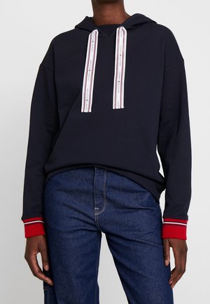 Osoba nosząca granatową bluzę z kapturem z białymi sznurkami oznaczonymi napisem "Tommy Hilfiger" oraz czerwonymi mankietami, połączoną z ciemnoniebieskimi jeansami.