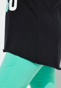Černé tričko s rovnými okraji, nošené přes světle tyrkysové slim-fit kalhoty. Tričko má grafický design v tyrkysové barvě.