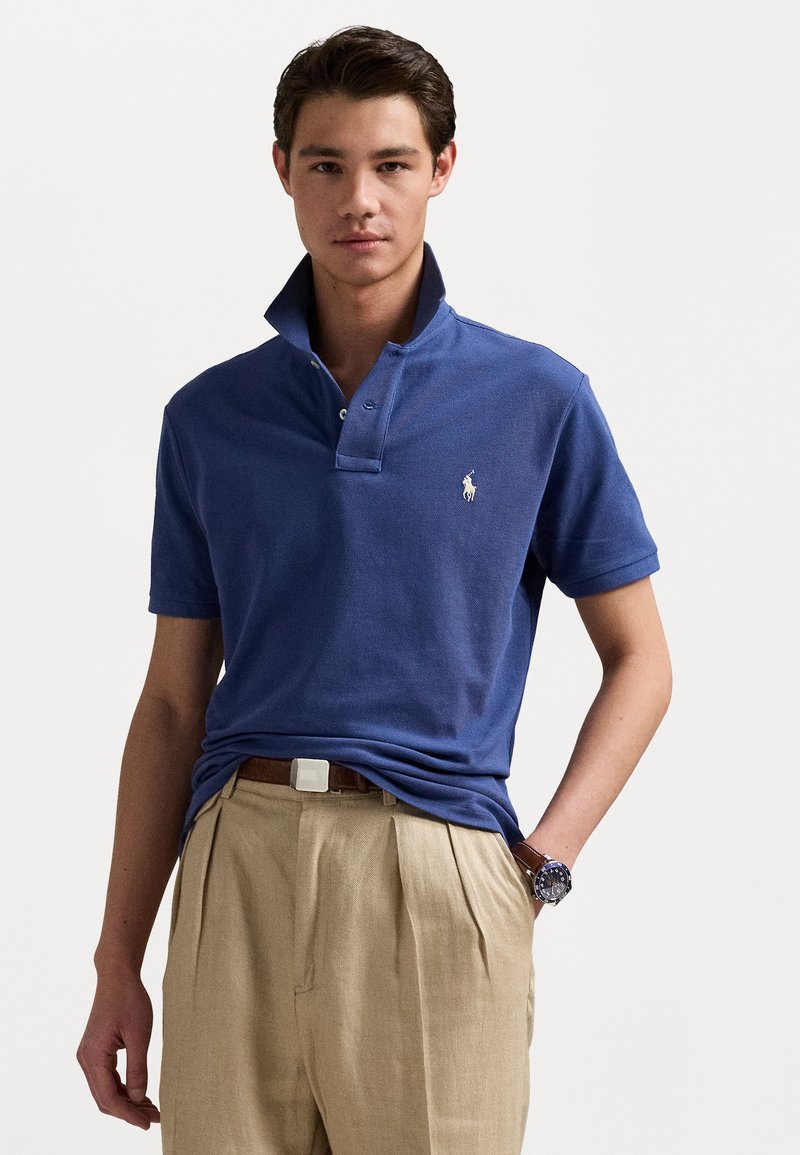 Dunkelblaues Poloshirt aus strukturiertem Stoff, mit zwei Knöpfen, erhöhter Kragen und kleinem weißen Logo auf der linken Brust. Brauner Gürtel und beigefarbene Hose.