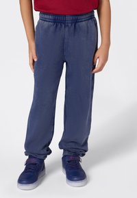 Kind in een blauwe joggingsbroek en blauwe sneakers met rood Champion-logo, staand tegen een effen lichte achtergrond.