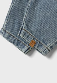 Denim blu sbiadito con taglio dritto, cuciture visibili e una piccola etichetta in pelle marrone attaccata all'orlo.