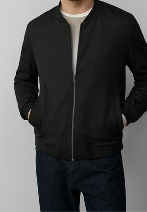 Homme portant une veste bomber noire zippée par-dessus une chemise beige, les mains dans les poches, assortie à un jean bleu foncé.