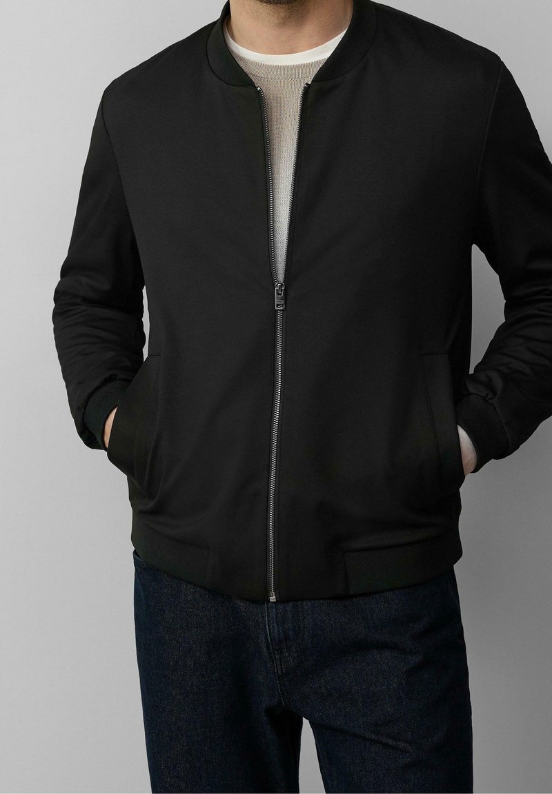 Mężczyzna ubrany w czarną kurtkę typu bomber zapinaną na zamek, na beżową koszulę, z rękami w kieszeniach, w połączeniu z ciemnoniebieskimi dżinsami.