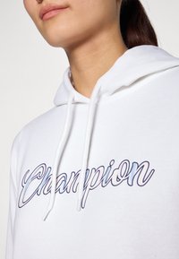 Felpa bianca con cappuccio, dotata di lacci, con il logo "Champion" in una scritta blu e rosa. Il materiale sembra morbido e leggermente testurizzato.