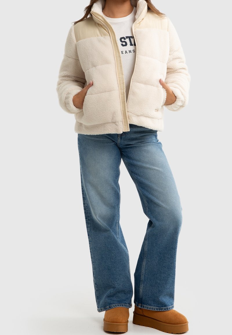 Veste en fausse fourrure crème avec un yoke en nylon beige clair, fermeture éclair, deux poches, associée à un jean large bleu et des bottes marron.
