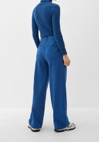 Blå långärmad tröja med hög krage i kombination med vida blå byxor, som har sidofickor och en mjuk yta. Skorna är klumpiga och mönstrade.