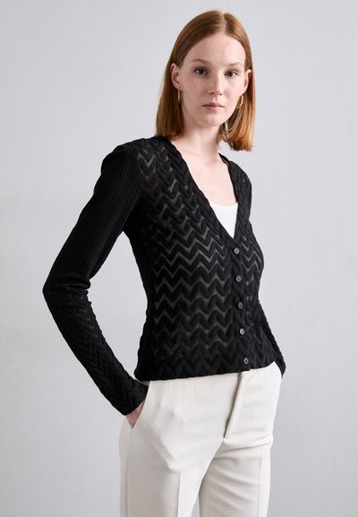 Cardigan noir avec une texture en chevrons, des manches longues et un décolleté en V profond, agrémenté de boutons et porté sur un haut blanc.