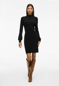 Abito a coste nere con scollo alto e maniche lunghe, abbinato a stivali in pelle marrone al ginocchio. Design semplice, silhouette aderente.