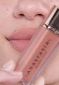 Płynna pomadka w przezroczystym opakowaniu z różowozłotą zakrętką. Usta są matowe i w delikatnym różowym odcieniu, mają gładką teksturę i subtelne konturowanie.