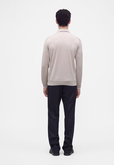 Homme debout, dos tourné, portant une chemise à manches longues gris clair, un pantalon bleu marine foncé et des chaussures noires sur un fond blanc uni.