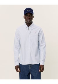 Lichtblauw gestreept overhemd met knopen en een klein rood logo. Draagt een marineblauwe pet met witte geborduurde tekst en casual denim jeans.