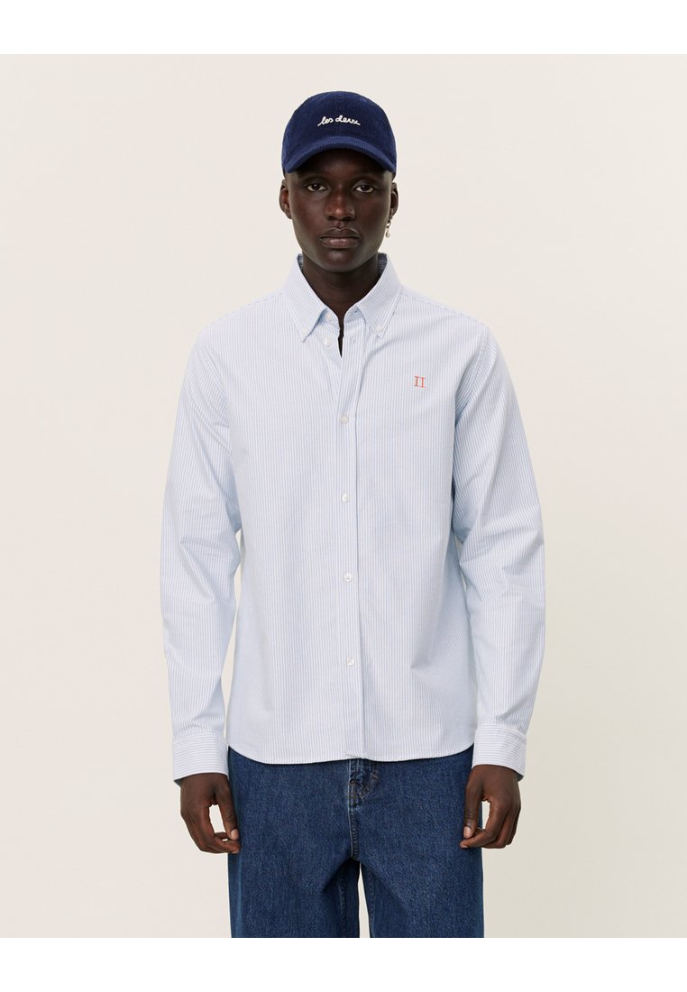 Lichtblauw gestreept overhemd met knopen en een klein rood logo. Draagt een marineblauwe pet met witte geborduurde tekst en casual denim jeans.