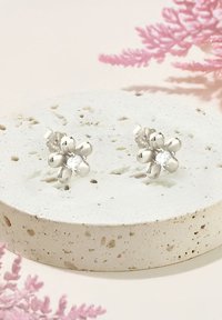 Silvriga blommformade stiftörhängen med en central klar ädelsten, visade på en texturerad vit stenyta, omgiven av mjuka rosa accenter.