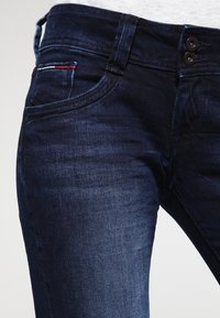 Donkerblauwe denim jeans met een smalle pasvorm, voorzien van een vijf-pocket ontwerp, sluiting met twee knopen en subtiele rode en witte stiksel details.