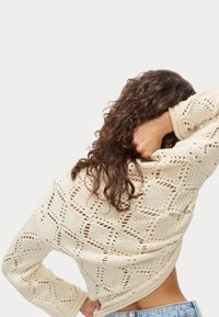 Crème gebreid sweater met een opengewerkt patroon en geometrische ontwerpen. Lange mouwen en een ontspannen pasvorm, gecombineerd met blauwe denim.