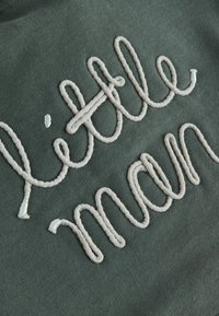 Grüner Stoff mit crèmeweißer, strukturiertem Stickerei, die "little man" in einer verspielten, geschwungenen Schriftart spellte, mit geschlungenem und gewundenem Design.