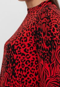 Vêtement rouge présentant un mélange de motifs léopard noirs et abstraits, avec un col montant et une coupe légèrement ample. Tissu texturé visible.