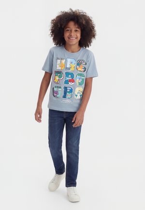 Uśmiechnięte dziecko z kręconymi włosami, ubrane w jasnoniebieski t-shirt z nadrukiem Pokémon, ciemne jeansy i białe trampki, idące do przodu na białym tle.