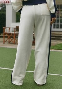 Balti pūkų sportinės kelnės su tamsiai mėlynomis šoninėmis juostomis, plataus kirpimo, su kišene gale. Tekstūruotas audinys ant žalios lauko paviršiaus.