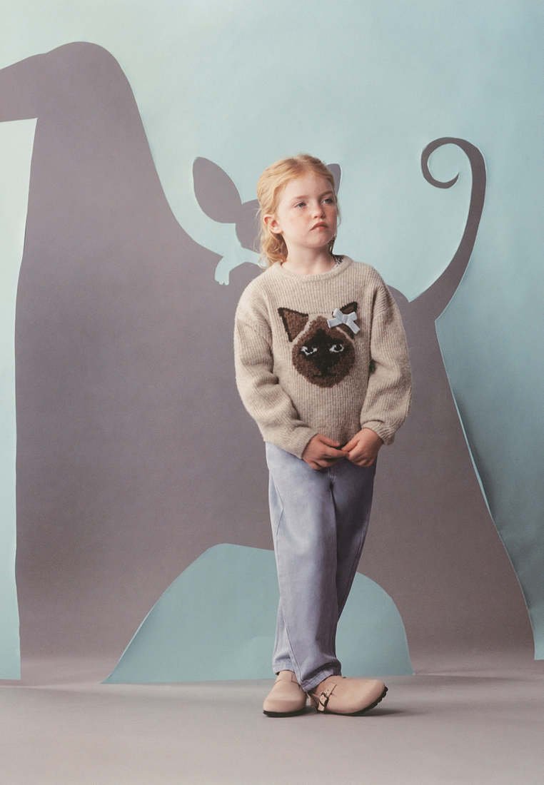 Jeune fille aux cheveux blonds portant un pull beige avec un motif visage de chat, un pantalon bleu clair et des chaussures beiges devant un décor en forme de chat.