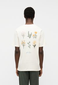 Άτομο που φοράει λευκό μπλουζάκι με έξι κεντητά άγρια λουλούδια σε δύο σειρές και το κείμενο "Les Deux" κάτω από τα σχέδια.