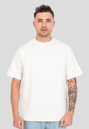 Mand med kort mørkt hår iført en enkel hvid kortærmet t-shirt og lyseblå jeans, der viser et tatoveringsærme på sin højre arm.