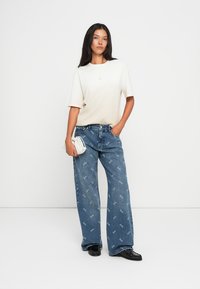 Valkoinen lyhythihainen t-paita yhdistettynä sinisiin farkkuihin, joissa on koko pinnan logo-printti, leveä lahje ja mustat kengät, joissa on teksturoitu pinta.