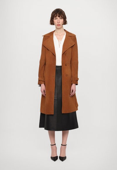 Cappotto lungo in suede marrone con ampi risvolti, design doppiopetto e polsini con cintura, indossato sopra una maglia bianca e una gonna in pelle nera.