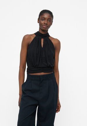 Γυναίκα με μαύρο τοπ χωρίς μανίκια και ψηλόμεσο, φαρδύ μαύρο παντελόνι, στέκεται μπροστά σε λευκό φόντο.