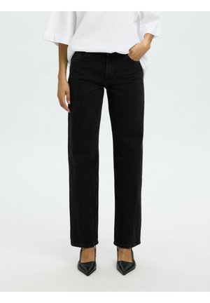 Osoba nosiaca čierne rifle so strihom rovnej nohy, bielu košeľu s dlhým rukávom a čierne lodičky s ostrou špičkou, stojaca s jednou rukou v vrecku.