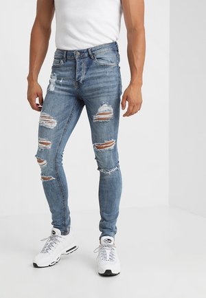 Mann trägt zerrissene blaue enge Jeans, weiße Turnschuhe und ein weißes T-Shirt, steht in einem minimalistischen weißen Raum.